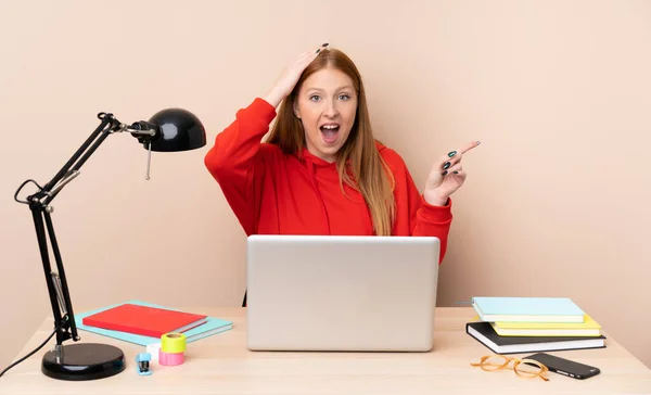 Young student woman in a workplace with a laptop surprised and pointing finger to the side