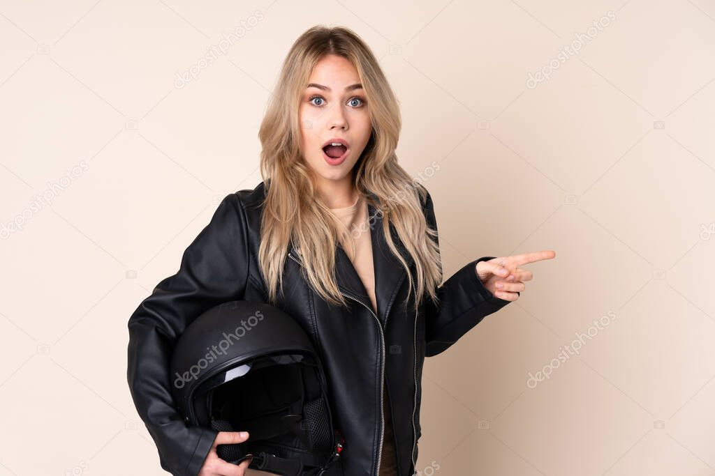Russian girl with a motorcycle helmet isolated on beige background surprised and pointing finger to the side