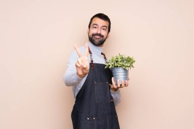 Arka planda bir bitkiyi tutan adam gülümsüyor ve zafer işareti gösteriyor.