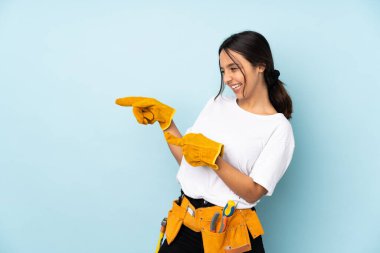 Genç elektrikçi kadın, mavi arka planda izole edilmiş bir şekilde parmak ucunda bir ürün sunuyor.