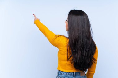 İzole edilmiş mavi arka plan üzerinde genç Kolombiyalı kız işaret parmağıyla işaret ediyor.