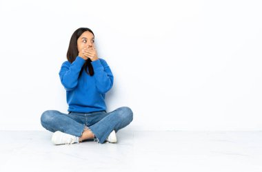 Genç melez kadın, beyaz arka planda izole bir şekilde yerde oturuyor. Ağzı örtüyor ve yan tarafa bakıyor.