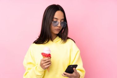 Genç esmer kız izole edilmiş pembe arka planda kornet dondurması tutuyor elinde kahve ve cep telefonu var.