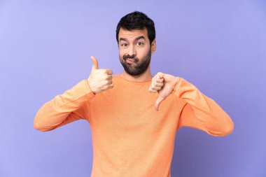 İzole edilmiş mor arka planda beyaz, yakışıklı bir adam iyiye-kötüye işaret yapıyor. Evet ya da hayır arasında kararsız