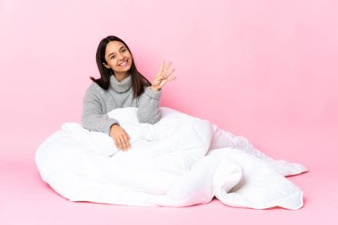 Genç melez kadın pijama giymiş yerde mutlu bir şekilde oturuyor ve parmaklarıyla dört tane sayıyor.