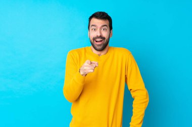 İzole edilmiş mavi arka plan üzerinde genç beyaz adam şaşırmış ve ön tarafı işaret ediyor.