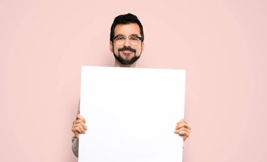 Handsome man with beard holding an empty placard over isolated pink background