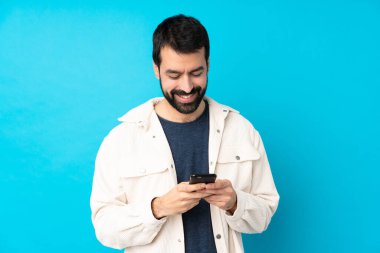 Mavi arka plan üzerinde beyaz kadife ceketli genç yakışıklı bir adam cep telefonuyla bir mesaj gönderiyor.