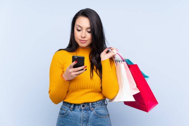 Genç Kolombiyalı bir kız, mavi arka planda, elinde alışveriş çantaları ve cep telefonuyla arkadaşına mesaj yazıyor.