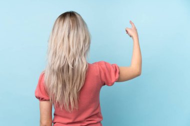 İzole edilmiş mavi arka planı olan genç kız işaret parmağıyla işaret ediyor.
