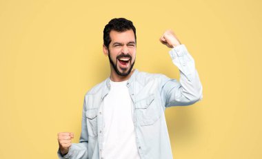 Sakallı yakışıklı adam izole edilmiş sarı arka plandaki zaferini kutluyor.