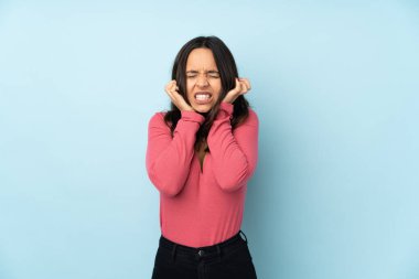 Mavi arka planda izole edilmiş genç bir ırk kadını hayal kırıklığına uğramış ve kulaklarını kapatmış.