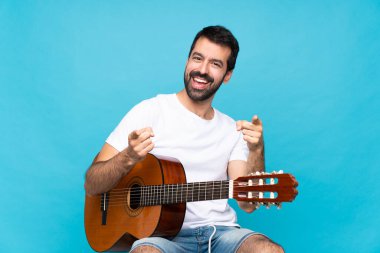 Mavi arka planda gitar çalan genç adam gülümserken seni işaret ediyor.