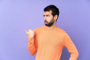 İzole edilmiş mor arka planı olan beyaz, yakışıklı bir adam mutsuz ve bir tarafı işaret ediyor.