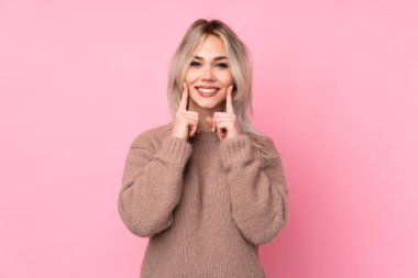 İzole edilmiş pembe arka planda süveter giyen genç sarışın kız mutlu ve hoş bir ifadeyle gülümsüyor.