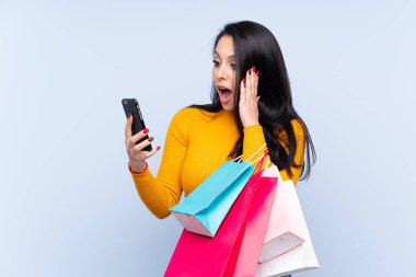 Genç Kolombiyalı bir kız, mavi arka planda, elinde alışveriş çantaları ve cep telefonuyla arkadaşına mesaj yazıyor.