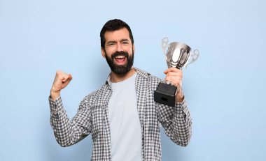 Sakallı yakışıklı bir adam mavi arka planda bir kupa tutuyor.