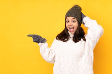 Sarı arka plan üzerinde kışlık şapkası olan genç kadın şaşırdı ve parmağını yana çevirdi.
