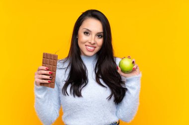 Kolombiyalı genç bir kız izole edilmiş sarı arka planda. Bir elinde çikolata tableti, diğer elinde elma tutuyor.