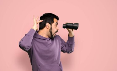 Handsome man with sweatshirt with binoculars over isolated pink background