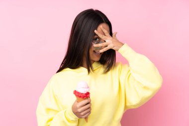 Genç esmer kız izole edilmiş pembe arka planda bir kornet dondurma tutuyor. Gözleri elleriyle kaplıyor ve gülümsüyor.