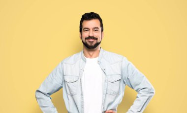 Kalçasında kollarıyla poz veren ve izole edilmiş sarı arka planda gülümseyen sakallı yakışıklı bir adam.