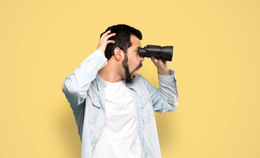 Handsome man with beard with binoculars over isolated yellow background