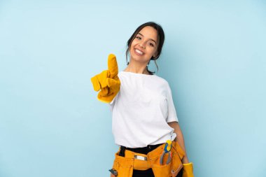 Mavi arka planda izole edilmiş genç elektrikçi kadın parmağını kaldırıyor ve gösteriyor