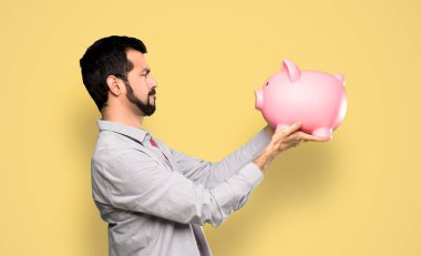 Handsome man with beard holding a piggybank over isolated yellow background