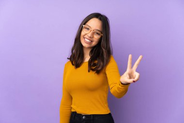 Mor arka planda izole edilmiş, gülümseyen ve zafer işareti gösteren genç melez bir kadın.