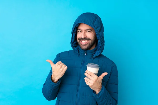 Man wearing winter jacket and holding a takeaway coffee over isolated blue background with thumbs up gesture and smiling