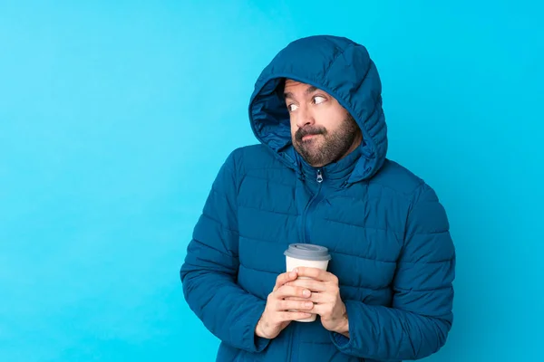 Man wearing winter jacket and holding a takeaway coffee over isolated blue background making doubts gesture looking side