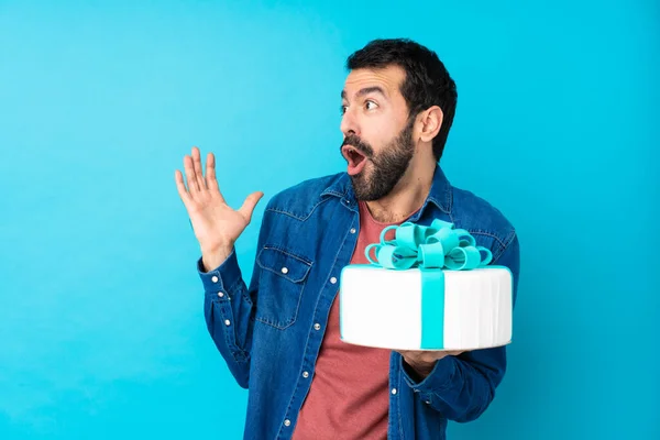 Young handsome man with a big cake over isolated blue background with surprise facial expression