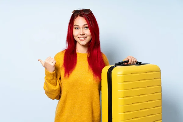 一个旅行女孩拿着一个蓝色背景的手提箱 指着旁边展示一个产品 — 图库照片