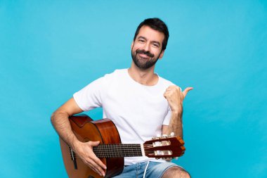 İzole edilmiş mavi arka planda gitar çalan genç bir adam. Baş parmakları havada ve gülümsüyor.