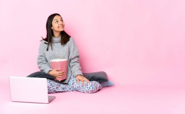 Genç melez kadın dizüstü bilgisayarda film izlerken patlamış mısır yiyor gülümserken yukarı bakıyor.