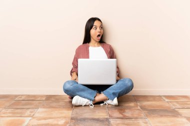 Dizüstü bilgisayarı olan genç melez bir kadın yerde oturuyor ve yan tarafa bakarken sürpriz jest yapıyor.