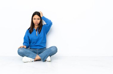 Genç melez kadın, beyaz arka planda izole bir şekilde yerde oturuyor. Hayal kırıklığına uğramış ve anlamamış bir ifadeyle.