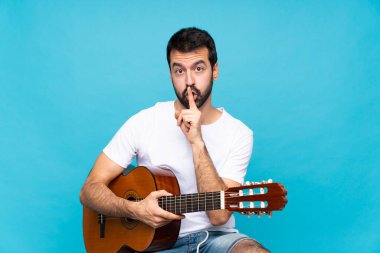 İzole edilmiş mavi arka planda gitarı olan genç bir adam sessizlik işareti gösteriyor. Parmağını ağzına sokuyor.
