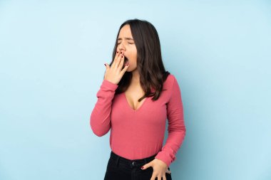 Genç melez kadın mavi arka planda izole edilmiş esniyor ve ağzı açık bir şekilde elleriyle ağzını kapatıyor.