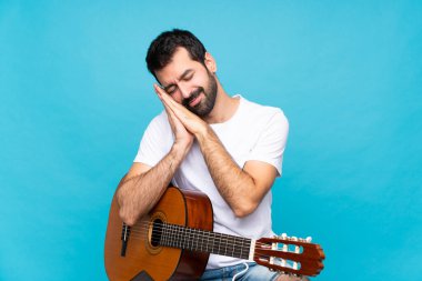 İzole edilmiş mavi arka planda gitarı olan genç bir adam uyku jesti yapıyor.