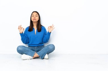 Zen pozisyonunda beyaz arka planda soyutlanmış bir şekilde yerde oturan genç melez bir kadın.