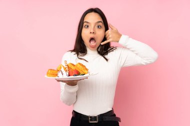 İzole edilmiş pembe arka planda waffle tutan esmer bir kadın telefon jesti yapıyor ve kuşkulanıyor.