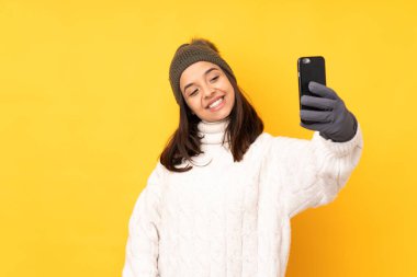 Sarı arka plan üzerinde kışlık şapkası olan genç bir kadın selfie çekiyor.