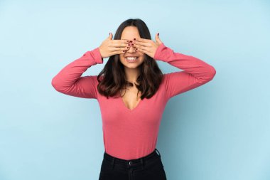 Mavi arka planda izole edilmiş genç melez kadın gözlerini elleriyle kapatıyor.
