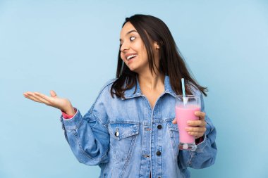 İzole edilmiş mavi arkaplanda çilekli milkshake 'i olan genç bir kadın iki eliyle kopyalama alanı tutuyor.