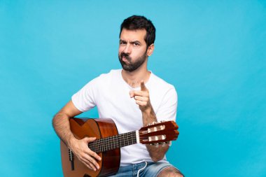 İzole edilmiş mavi arka planda gitar çalan genç adam hayal kırıklığına uğramış ve ön tarafı işaret ediyor.