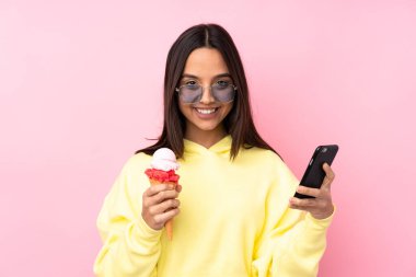 Genç esmer kız izole edilmiş pembe arka planda kornet dondurması tutuyor elinde kahve ve cep telefonu var.