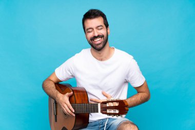 İzole edilmiş mavi arka planda gitar çalan genç adam çok gülümsüyor.