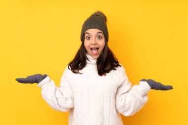 Kışlık şapkalı genç bir kadın izole edilmiş sarı bir arka plan ve şok olmuş bir yüz ifadesi.